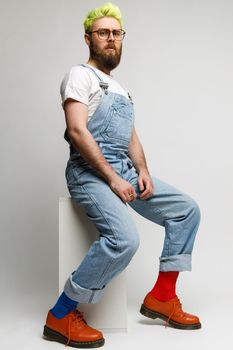 Full length photo of confident concentrated man wearing jacket, denim overalls and stylish red shoes, looking at camera with serious expression. Indoor studio shot isolated over gray background.