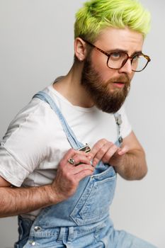 Side view of serious bearded model with green hair guy looking away with concentrated expression, fastening clasp on his trendy denim overalls. Indoor studio shot isolated over gray background.