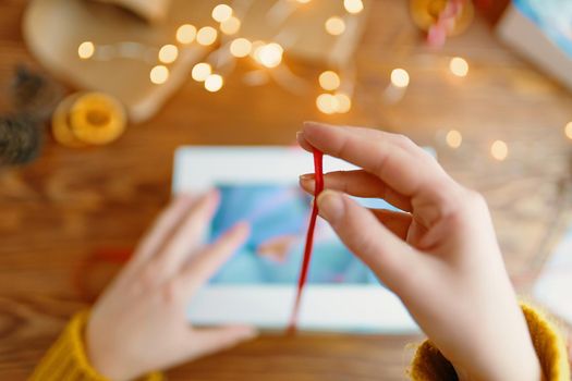Close-up of woman's hand in knitted sweater holding red ribbon. Gift box and Christmas decorations on wooden table. New Year's composition.