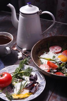 Fried eggs with young garlic stems, greenery, basil, rosemary and tomatoes on frying pan with a cup of coffe and coffe pot, selective focus.