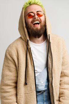 Handsome young adult bearded model man wearing beige jacket looking at camera and laughing out loud, expressing positive and optimism. Indoor studio shot isolated over gray background.