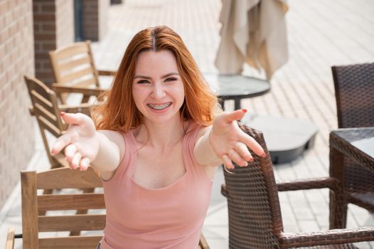 Young red-haired woman sits in a street cafe and stretches her arms to hug