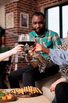 Cheerful people toasting wineglasses while celebrating birthday at home. Diverse group of close friends at wine party clinking glasses while enjoying fun time together in living room.