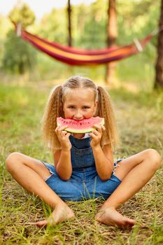 Funny girl eating watermelon in the park. Kid eat fruit outdoors. Healthy snack for children. Little girl playing in the forest biting a slice of water melon