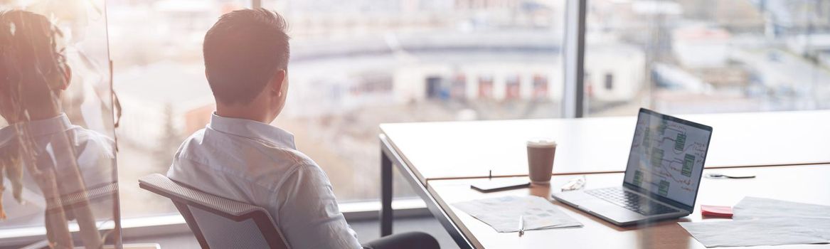 Focused businessman working on laptop and looking out the window at her workplace at modern office.