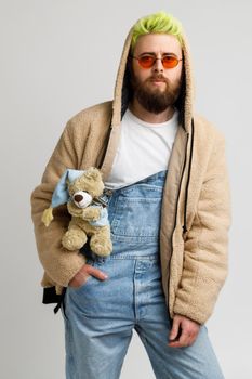 Young attractive bearded man model wearing stylish jacket and denim overalls, keeping hand in pocket, looks at camera with pensive expression. Indoor studio shot isolated over gray background.