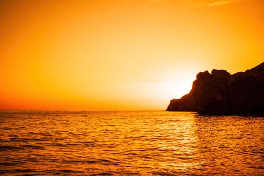 A red burning sunset with the silhouette of a cliff over the sea. Abstract nature summer or spring ocean sea background. Small waves on golden warm water surface with bokeh lights from sun.