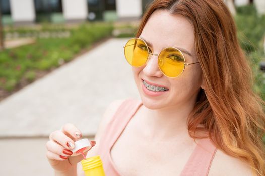 Young red-haired woman blowing soap bubbles outdoors. Girl in yellow sunglasses and braces