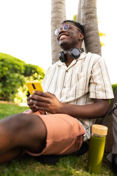 Happy black man relaxing outdoors using mobile phone. College student on campus. Vertical image. Technology and healthy lifestyle concept.