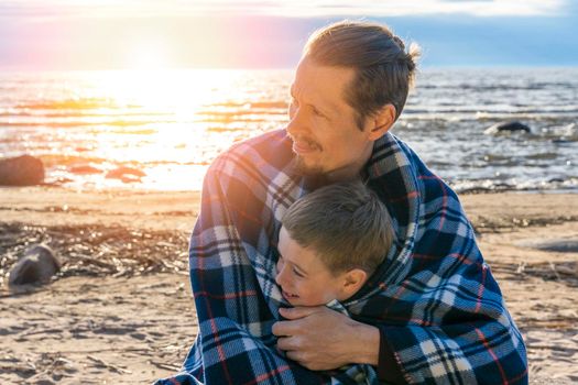 a father and his preschooler son are relaxing on the beach wrapped in a plaid plaid. the father hugs his son. parental love. father's day