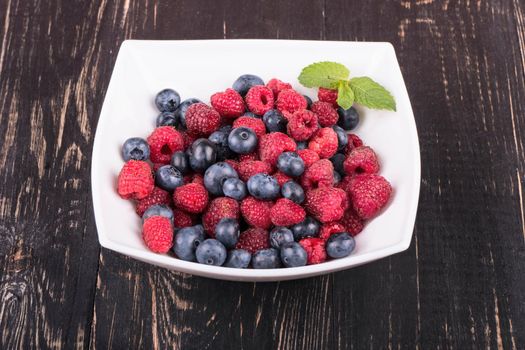 Raspberries and blueberries in a white bowl on a wooden background