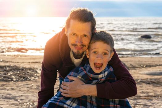 a father and his preschooler son are relaxing on the beach wrapped in a plaid plaid from the wind. the father hugs his son. parental love. father's day