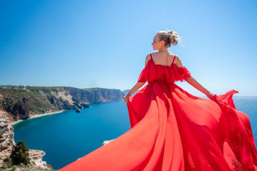 Blonde with long hair on a sunny seashore in a red flowing dress, back view, silk fabric waving in the wind. Against the backdrop of the blue sky and mountains on the seashore