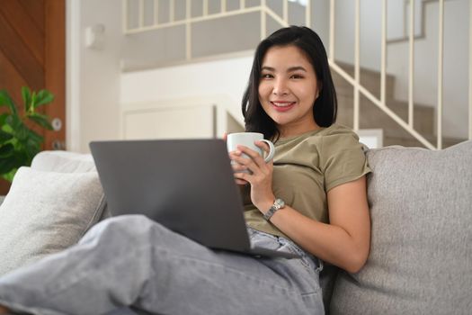 Casual young woman drinking hot tea and using laptop on couch in bright living room.