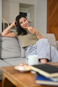 Smiling young woman eating donut and watching television on comfortable sofa at home.