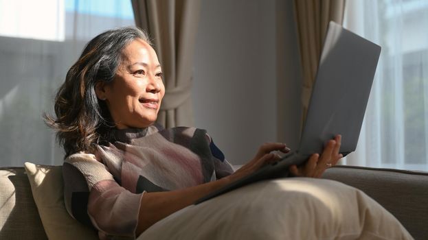 Pleasant mature woman surfing internet, reading online news on laptop while relaxing on couch. Elderly technology concept.