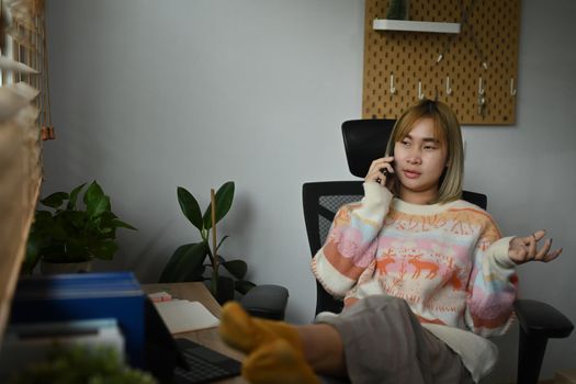 Asian woman sitting in home office talking on mobile phone.