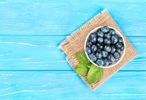 White bowl with fresh blueberries, mint leaves on an empty wooden background, top view