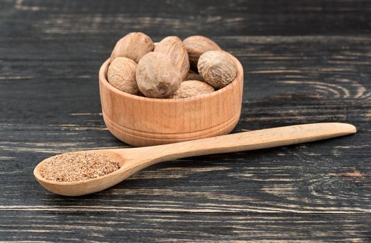 Nutmeg powder in a spoon with a bowl of nuts on wooden background