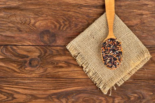 Black rice in spoon on burlap and empty wooden background