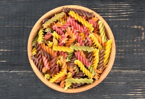 Colored pasta fusilli in a bowl on wooden background, top view