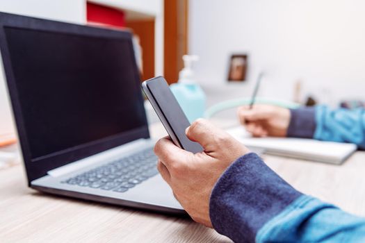 detail of the hand of a man holding a smartphone, out of focus is a computer and a hand sanitizer gel, work at home concept due to the coronavirus pandemic
