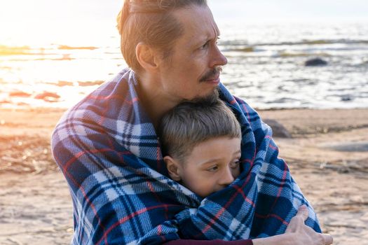 a father and his preschooler son are relaxing on the beach wrapped in a plaid plaid. the father hugs his son. parental love. father's day