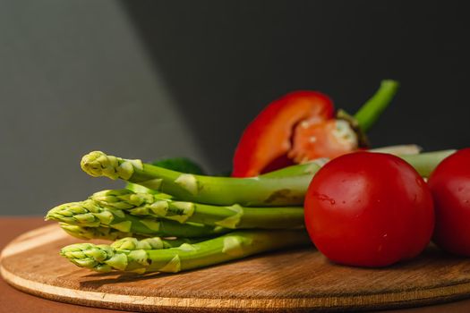 Vegetables lie on a wooden board: tomatoes, asparagus, cucumbers, red bell peppers. brown, dark gray background. place for text