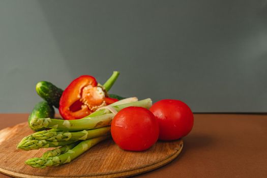 Vegetables lie on a wooden board: tomatoes, asparagus, cucumbers, red bell peppers. brown, dark gray background. place for text