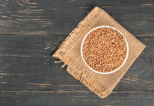White bowl with raw buckwheat on burlap and empty wooden background