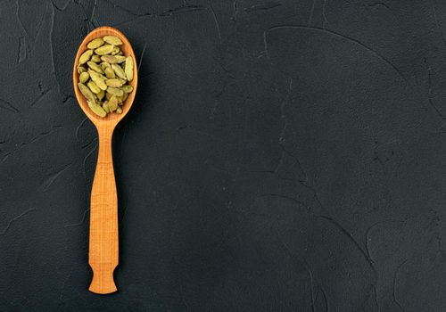 Green cardamom in wooden spoon on dark background, top view