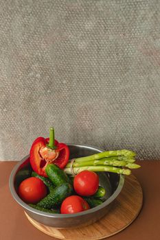 Vegetables lie in a metal bowl: tomatoes, asparagus, cucumbers, red bell peppers . on a wooden board and brown background. back gray background. place for text.