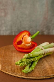 branches of fresh green asparagus on a wooden board, red big pepper on the background. brown background. Basic trend concept with copy space