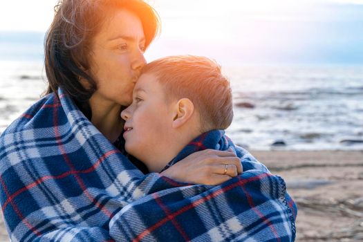 a happy mother and her preschooler son are relaxing on the beach wrapped in a plaid plaid from the wind in the light of the setting sun. parental love. family ties. Mother's Day