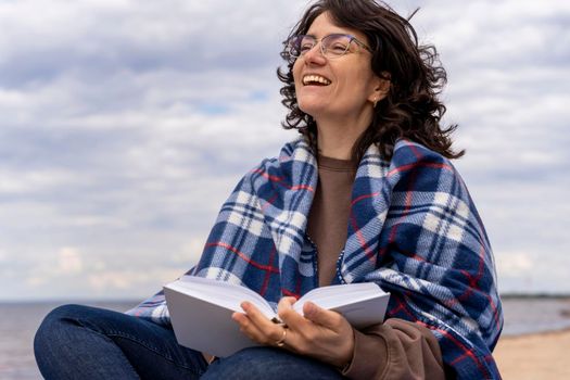 A cheerful woman with glasses reads a book on the seashore covered with a blanket. Cozy picnic and relaxing outdoor recreation with a book, enjoying life and fresh air