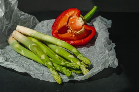 fresh green asparagus and half of red sweet, large pepper lie on white parchment on a dark gray background. proper nutrition. Lifestyle. place for text