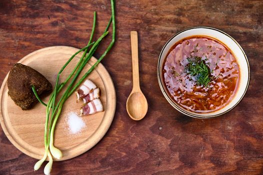 High angle view of a delicious, tasty national Ukrainian borscht and wooden board with green onion, salt, bacon and bread on wooden rustic table. Slavic traditional dish. Copy ad space . Flat lay.