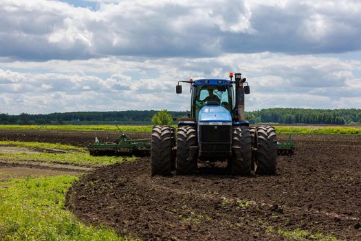 Blue New Holland tractor with double wheels pulling disc harrow with roller basket at hot sunny day in Tula, Russia - June 4, 2022