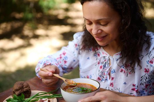Close-up of a middle-aged charming multi-ethnic woman eating delicious traditional Ukrainian borscht - beetroot soup with whole grain rye bread and green onion outdoors. Slavic cooking