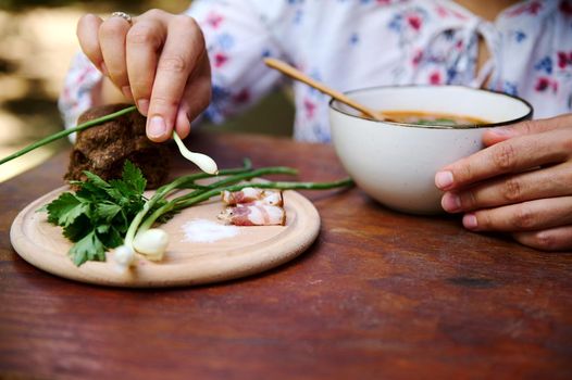 Close-up of a woman holding green onion while tasting Ukrainian national Borscht on a served rustic wooden table. Slavic traditional culinary. Red beetroot soup, sliced bacon, parsley on cutting board