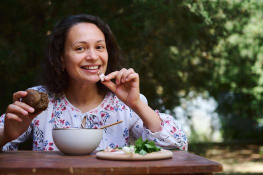 African American beautiful delightful woman sitting at rustic wooden dining table, having lunch outdoors, eating traditional ukrainian borscht with bacon slice and rye bread, smiling looking at camera