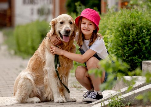 Preteen girl wearing hat hugging golden retriever dog sitting outdoors in summertime. Pretty kid petting fluffy doggy pet in city