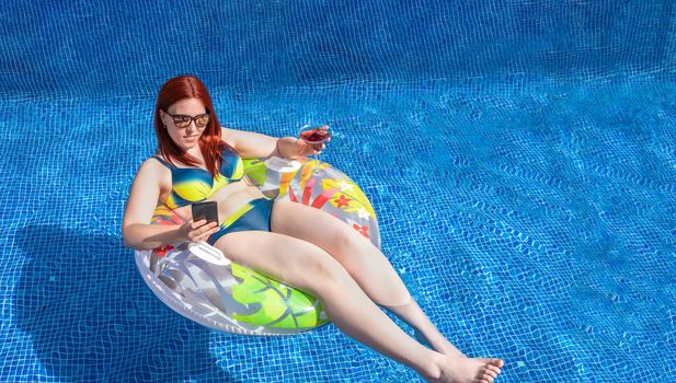 young red-haired girl, looking at her smartphone floating on a ring float inside the pool, while holding a soft drink. young girl on holiday relaxing. concept holiday and travel. outdoor garden, natural sunlight.