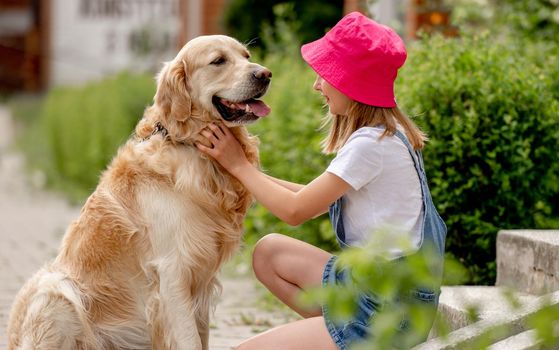 Preteen girl wearing hat hugging golden retriever dog sitting outdoors in summertime. Pretty kid petting fluffy doggy pet in city