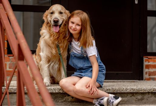Preteen girl wearing jeans dress with golden retriever dog sitting outdoors in summertime. Pretty kid hugging purebred fluffy doggy pet in city