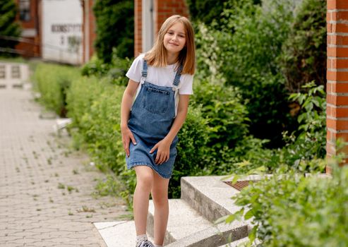 Preteen girl wearing jeans dress in city in summertime smiling. Beautiful female kid looking at camera in sunny day