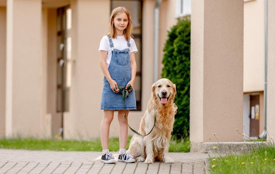Preteen girl wearing jeans dress with golden retriever dog outdoors in summertime looking at camera. Pretty kid with purebred fluffy doggy pet in city