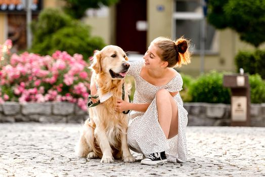 Girl in beautiful dress hugging golden retriever dog at street in summer sunny day. Pretty teenager petting purebred doggy pet in city with ancient architecture