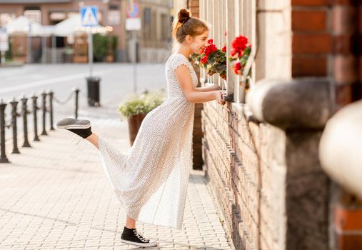 Beautiful girl in long dress sniffing red flowers at street in city with ancient architecture. Pretty female teenager model outdoors in summer time