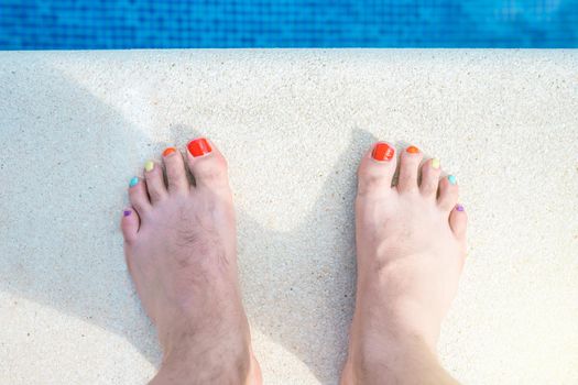 feet of a hairy man with his nails painted in LGBTIQ colours. bare feet at the edge of a swimming pool. concept of holidays and diversity. outdoors, natural light, summer days.